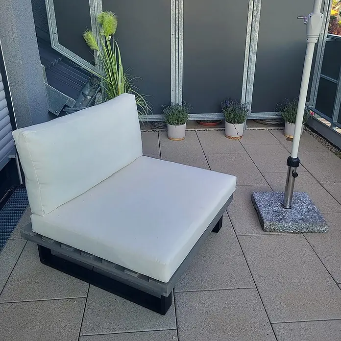 Mendler Garten-Loungesessel HWC-H54 - 1 Stk., Holz, Grau, Polster cremeweiß, B x T x H: 82 x 82 x 75 cm - Ein Outdoor-Loungesessel mit weißen Kissen steht auf einer gefliesten Terrasse. Er hat einen grau-schwarzen Palettenunterbau für Entspannung.