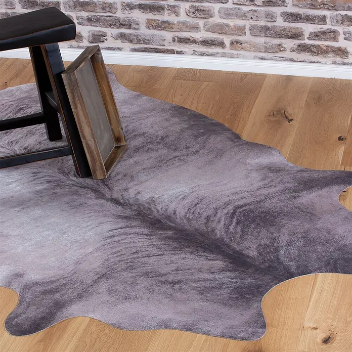 Kayoom Kunstfell Sanari 203 - Grau, 190 x 155 cm, 100 % Polyester - Grauer Kunstfell-Kuhfellteppich mit meliertem Muster auf hellem Holzboden unter einem schwarzen Hocker.