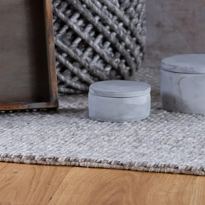 Kayoom Fransenteppich Pixy 525 - Grau, 170 x 120 cm, 60 % Wolle, 40 % Viskose - Ein strukturierter, hellgrauer Webteppich liegt auf einem Holzfußboden, umgeben von dekorativen Betonbehältern und einem Holztablett.
