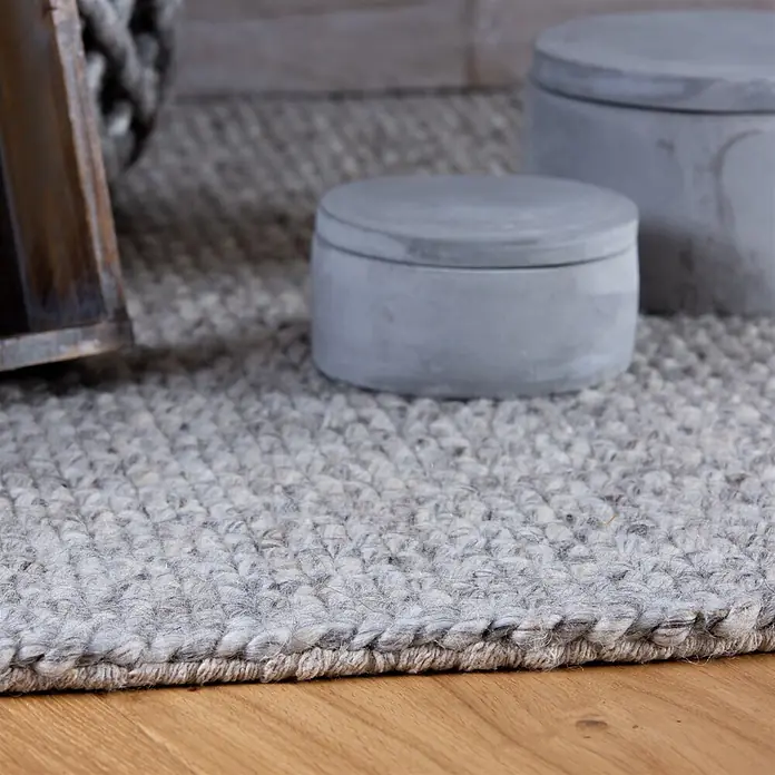 Kayoom Fransenteppich Pixy 525 - Grau, 170 x 120 cm, 60 % Wolle, 40 % Viskose - Hellgrauer, texturierter Webteppich auf Holzboden mit dekorativen Betonbehältern im Hintergrund.