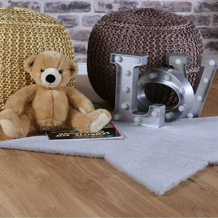 Kayoom Kinderteppich Lille 868 - Grau, 86 x 86 cm, 100 % Polyester - Ein weicher, hellgrauer Kunstfellteppich auf einem Holzfußboden, mit einem Teddybär und Dekobuchstaben.