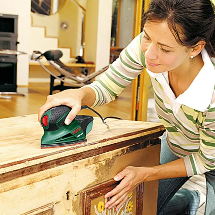 Bosch Easy Lijadora multifunción eléctrica  PSM 80 A - 80 W, Número de oscilaciones marcha en vacío: 22.000 r.p.m., Diámetro circuito oscilante: 1,4 mm - Mujer lijando un cofre con una lijadora multifunción verde Bosch, eliminando pintura vieja y preparando la superficie.