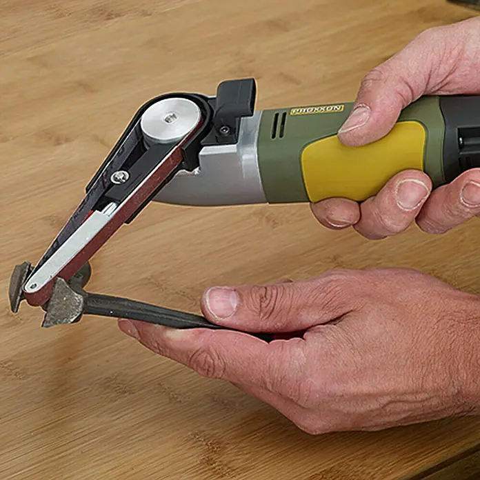 Proxxon Micromot Elektro-Bandschleifer BS/E - 80 W - Nahaufnahme des PROXXON Bandschleifers beim Bearbeiten eines Metallstücks auf einer Holzoberfläche.
