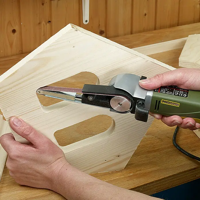 Proxxon Micromot Elektro-Bandschleifer BS/E - 80 W - Nahaufnahme einer Person, die eine Holzkonstruktion mit einem grünen Bandschleifer bearbeitet.