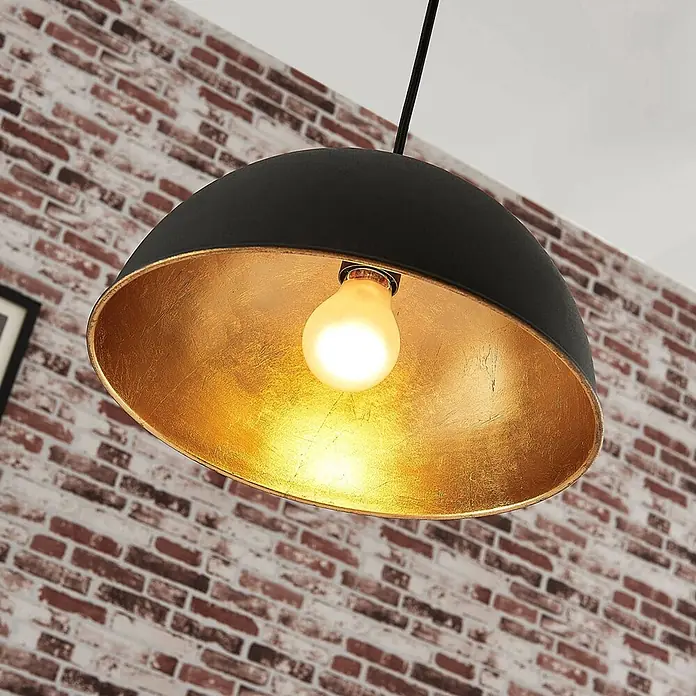 Lindby Pendelleuchte Leya - schwarz, Schwarz, gold, E27 - Schwarze Pendelleuchte mit goldener Innenseite und warmer Glühbirne vor einer Ziegelwand.