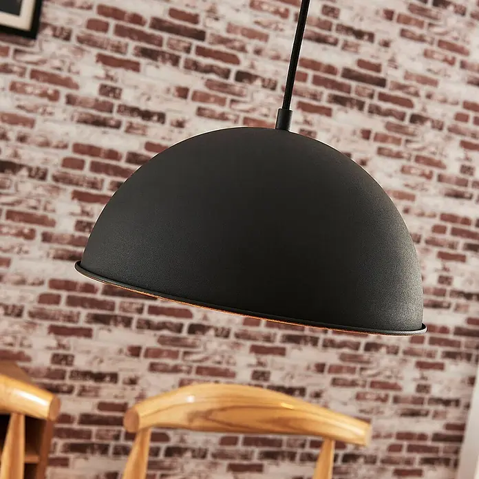 Lindby Pendelleuchte Leya - schwarz, Schwarz, gold, E27 - Schwarze, matte Hängeleuchte mit kuppelförmigem Schirm, vor einer Ziegelwand und Holzstühlen.