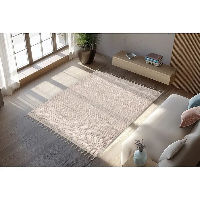 Kayoom Fransenteppich Reef 125 - Beige, 170 x 120 cm, 100 % Polypropylen - Ein heller beiger Teppich mit geometrischem Muster und Quasten, auf einem Holzfußboden in einem sonnigen Wohnzimmer mit Sofa.