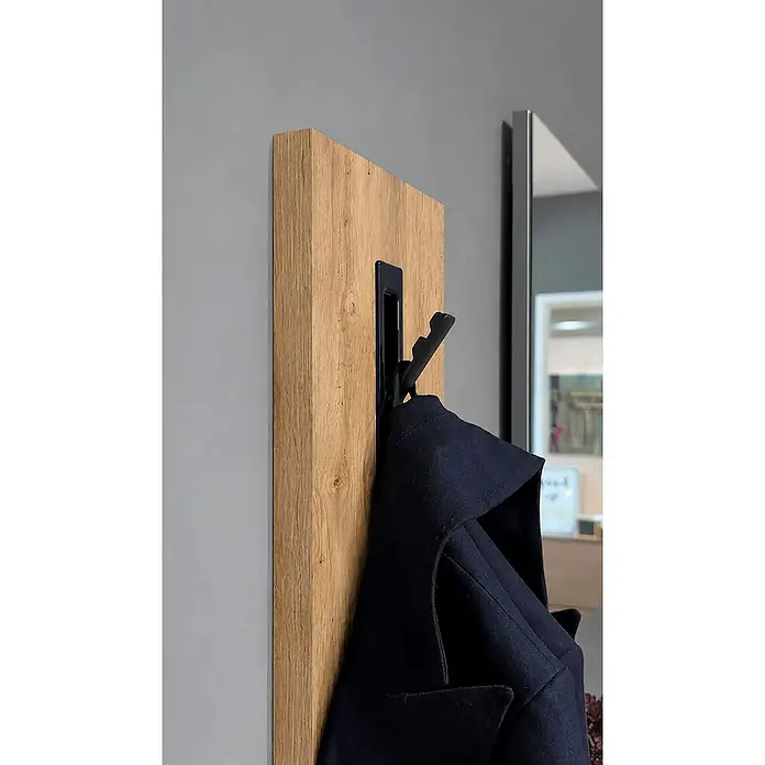 Schildmeyer Saga Garderobenpaneel - L x B x H: 2,5 x 39 x 109 cm, Holzwerkstoff, Eiche - Helle Holz-Wandpaneel mit schwarzem, ausklappbarem Haken, der einen dunklen Mantel hält, neben einem Spiegel.