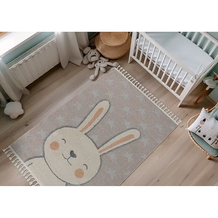 Kayoom Kinderteppich Modena 415 - Beige, Durchmesser: 133 cm, 100 % Polypropylen - Beiger Kinderteppich mit großem weißem Hasenmotiv, hellblauen Sternen und Quasten, auf Holzboden.