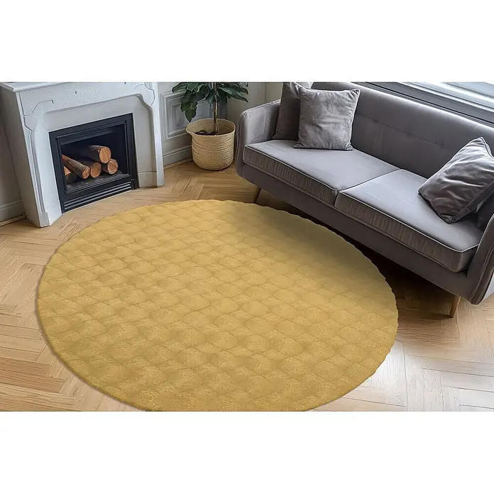 Kayoom Fellteppich Royal Glow 825 - Honig, Durchmesser: 120 cm, 80 % Polyester, 20 % Baumwolle - Runder, gelber Strukturteppich in einem Wohnzimmer, teilweise unter einem grauen Sofa und neben einem Kamin.