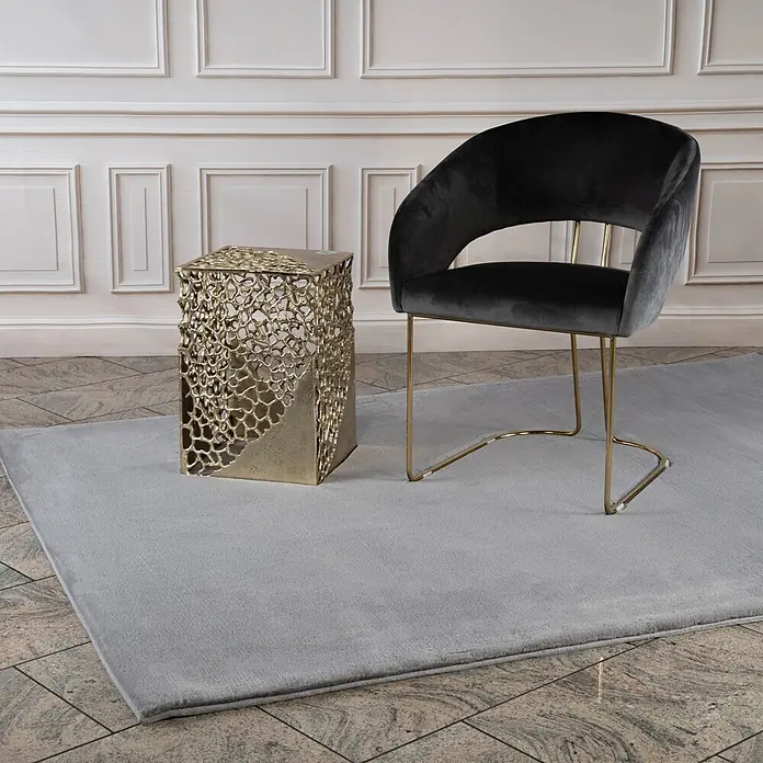 Kayoom Fellteppich Cross 425 - Silber, 170 x 120 cm, 100 % Polyester - Ein weicher, hellgrauer Teppich, teilweise bedeckt von einem goldenen Beistelltisch und einem schwarzen Samtsessel.