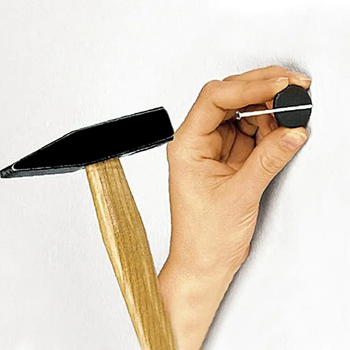 Alpha Tools Schlosserhammer - 100 g, Eschenholz - Nahaufnahme eines Hammers und einer Hand, die einen Nagel in eine Wand schlägt. Das Werkzeug ist ideal für Reparaturarbeiten im Haus.