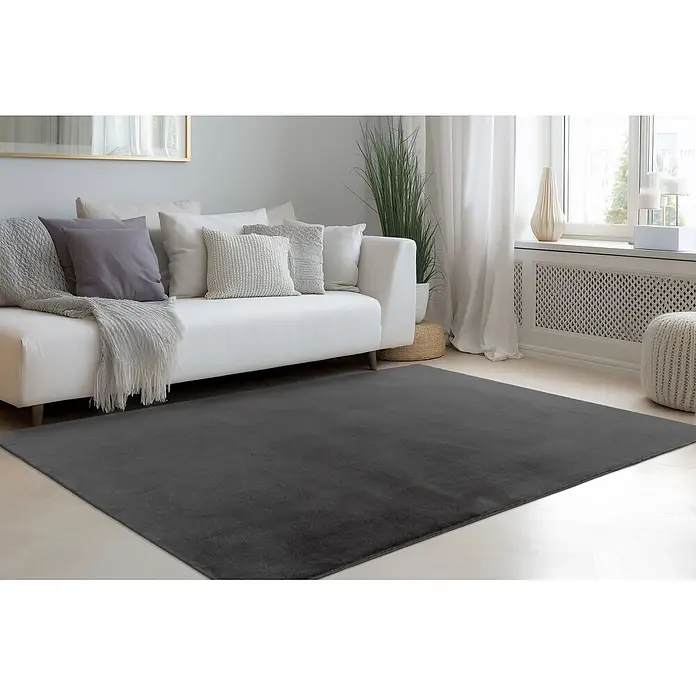 Kayoom Fellteppich Emperor 525 - Graphit, 150 x 80 cm, 100 % Polyester - Dunkelgrauer Teppich auf hellem Holzboden, teilweise unter einem weißen Sofa in einem Wohnzimmer.