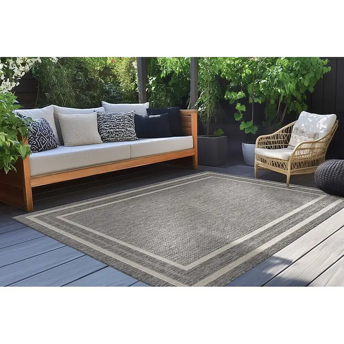 Kayoom Outdoor-Teppich Nibru 325 - Silber, 110 x 60 cm, 100 % Polypropylen - Grauer Outdoor-Teppich mit hellem Rand auf einer Terrasse mit Gartenmöbeln.