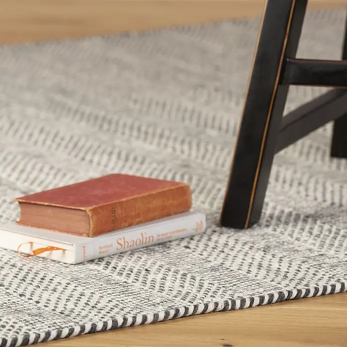 Kayoom Wollteppich Bombay 343 - Silber, 200 x 140 cm, 100 % Wolle - Gewebter grau-weißer Zickzack-Teppich auf Holzboden, mit Büchern und einem schwarzen Stuhlbein.