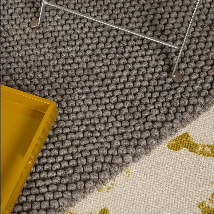 Kayoom Wollteppich Attic 590 - Taupe, 170 x 120 cm, 50 % Wolle, 50 % Viskose - Teppich mit grauen Filz-Wollkugeln und einem gewebten cremefarbenen Bereich mit gelben Mustern, teilweise unter einem Metallbein.