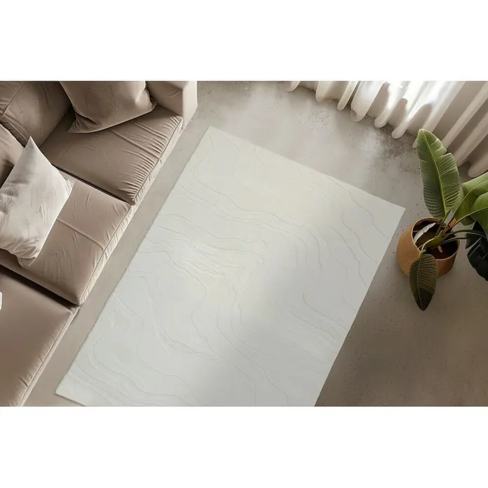 Kayoom Kurzflorteppich Cosmic 525 - Weiß, 230 x 160 cm, 85 % Polyester, 15 % Wolle - Ein cremefarbener Teppich mit subtilen Wellenmustern liegt auf einem Betonboden in einem Wohnzimmer neben einem beigefarbenen Sofa und einer Topfpflanze.