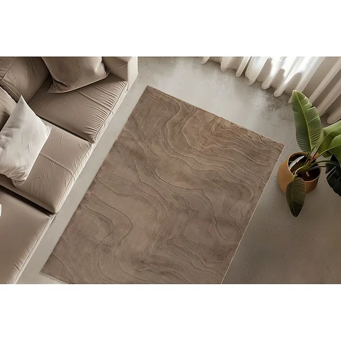 Kayoom Kurzflorteppich Cosmic 525 - Taupe, 230 x 160 cm, 85 % Polyester, 15 % Wolle - Ein rechteckiger beigefarbener Teppich mit welligem, strukturiertem Muster auf einem Betonboden neben einem beigen Sofa und einer Topfpflanze.