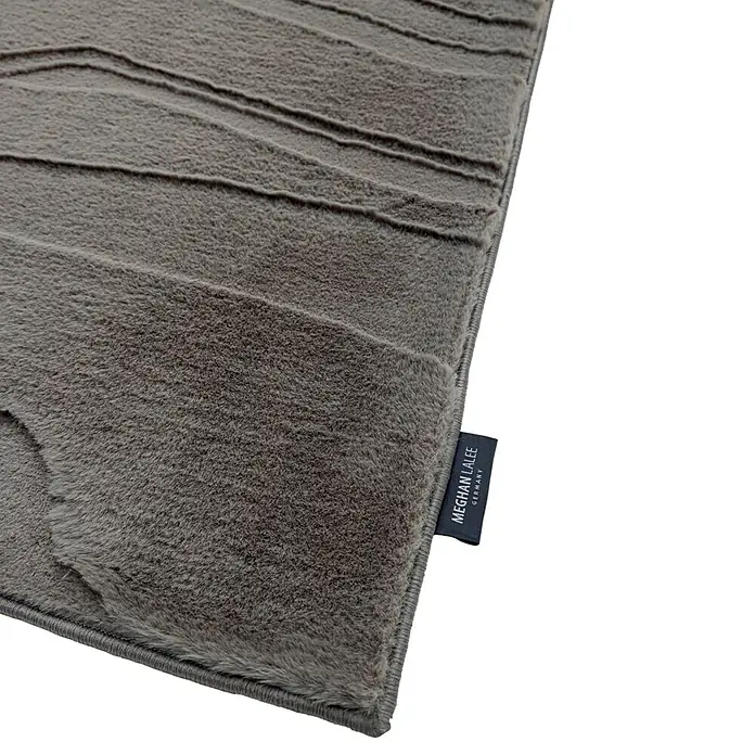 Kayoom Kurzflorteppich Cosmic 525 - Grau, 230 x 160 cm, 85 % Polyester, 15 % Wolle - Nahaufnahme eines grauen Plüschteppichs mit dezentem Wellenmuster und „MEGHAN LALEE GERMANY“-Etikett.