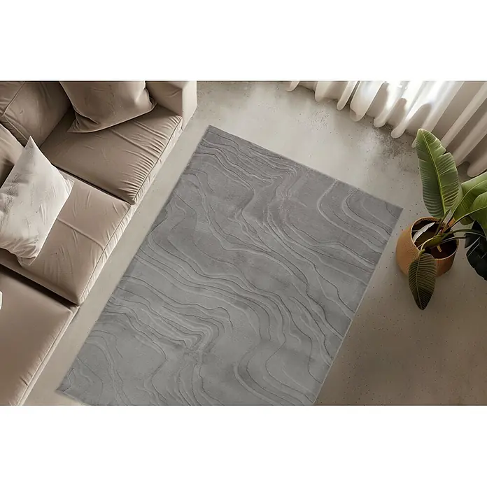 Kayoom Kurzflorteppich Cosmic 525 - Grau, 230 x 160 cm, 85 % Polyester, 15 % Wolle - Grauer Teppich mit Wellenmuster auf Betonboden, neben beigem Sofa und Pflanze.