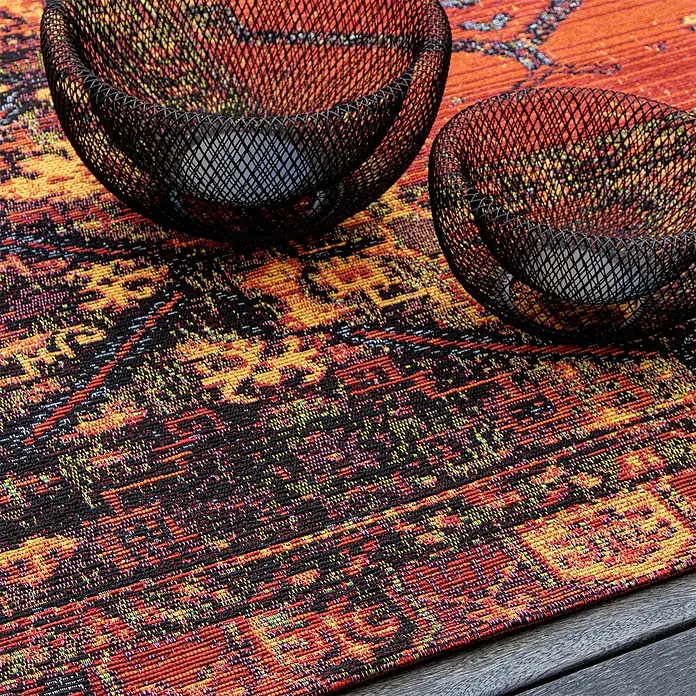 Kayoom Outdoor-Teppich Goliath 650 - Multi, 150 x 80 cm, 76 % Polypropylen, 24 % Polyester - Zwei schwarze Netzschalen auf einer lebhaften, gemusterten Platzmatte auf einem dunklen Holztisch.