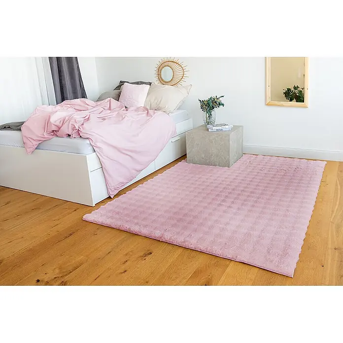 Kayoom Fellteppich Hailey 125 - Rosa, 290 x 200 cm, 100 % Polyester - Ein rechteckiger, flauschiger rosa Teppich mit subtiler Mustertextur liegt auf einem Holzboden neben einem Bett.