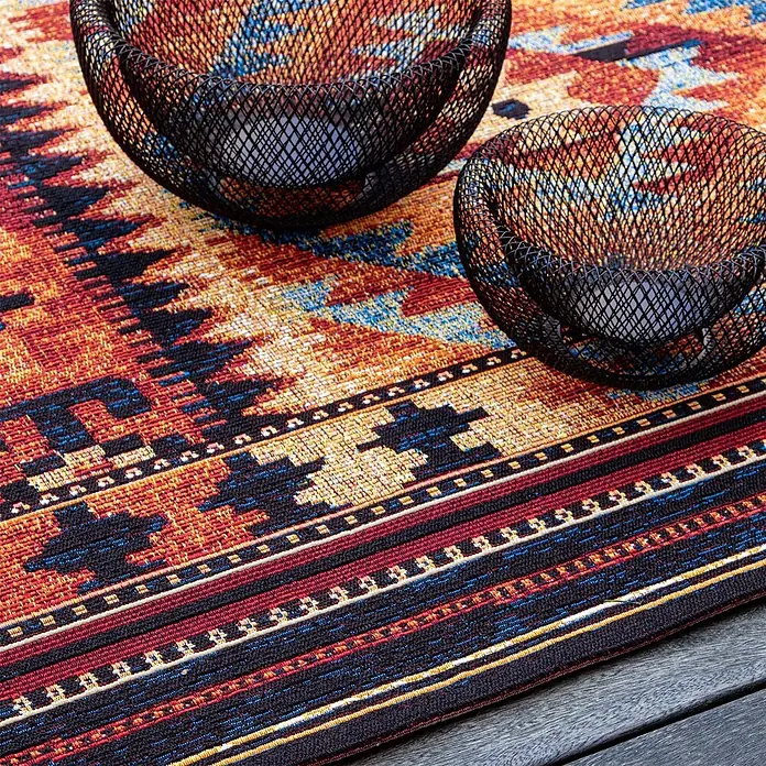 Kayoom Outdoor-Teppich Goliath 651 - Multi, 290 x 200 cm, 76 % Polypropylen, 24 % Polyester - Zwei schwarze Teelichthalter aus Metallgeflecht stehen auf einem bunten, gemusterten Stoffuntersetzer auf einer Holzoberfläche.