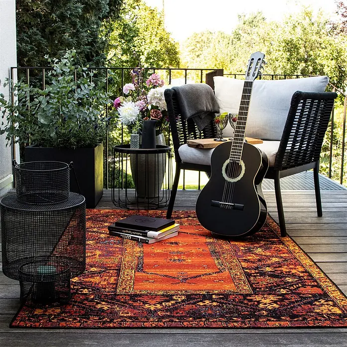 Kayoom Outdoor-Teppich Goliath 650 - Multi, 150 x 80 cm, 76 % Polypropylen, 24 % Polyester - Ein orange-schwarz gemusterter Outdoor-Teppich auf Holzbalkonboden, der einen gemütlichen Sitzbereich aufwertet.