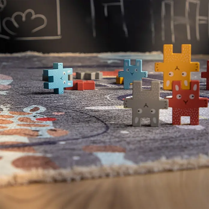 Kayoom Kinderteppich Joanna 610 - Spacegrau, 170 x 115 cm, 70 % Baumwolle, 30 % Polyester - Bunte Holz-Puzzlefiguren in Hasenform stehen auf einer gemusterten Spielmatte, was ein Kinderspiel andeutet.