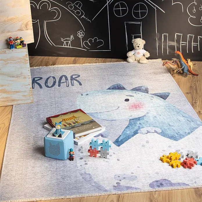 Kayoom Kinderteppich Joanna 626 - Multi, 170 x 115 cm, 70 % Baumwolle, 30 % Polyester - Kinderteppich mit blauer Dinosaurier-Illustration und „ROAR“-Text, auf Holzfußboden in Spielzimmer mit Spielzeug.