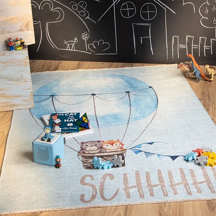 Kayoom Kinderteppich Joanna 611 - Multi, 170 x 115 cm, 70 % Baumwolle, 30 % Polyester - Ein hellblauer Teppich mit Heißluftballon, zwei Tieren im Korb und SCHHHH! Text, in einem Kinderzimmer mit Spielzeug ausgelegt.