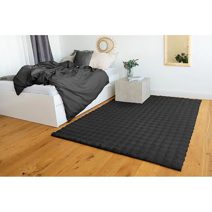 Kayoom Fellteppich Hailey 125 - Schwarz, 290 x 200 cm, 100 % Polyester - Ein dunkelgrauer, plüschiger Teppich mit Textur liegt auf Holzboden in einem Schlafzimmer neben einem Bett.