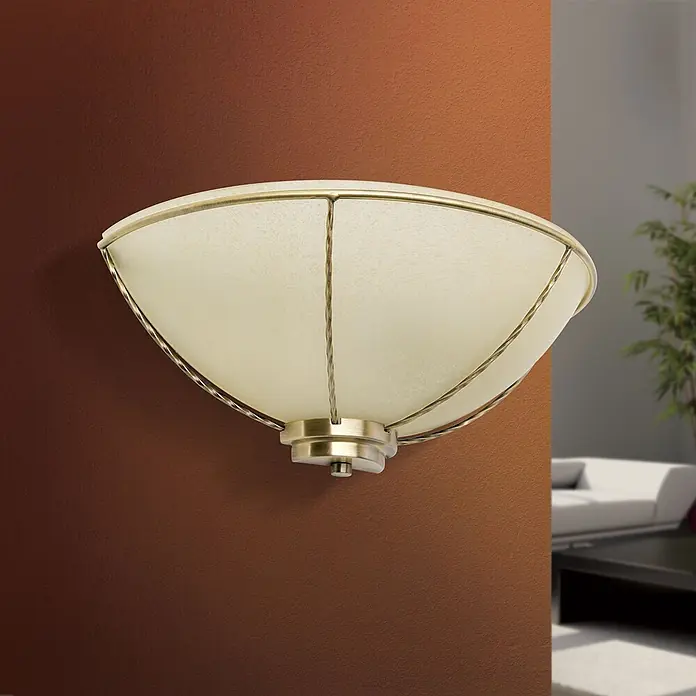 Orion Leuchten Wandleuchte - 60 W, L x B x H: 16 x 31 x 13 cm, E27 - Eine verzierte Wandleuchte mit creme-farbenem strukturiertem Glasschirm und antik-messingfarbenem Metallrahmen, an einer braunen Wand im Wohnzimmer montiert.