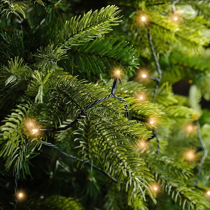 Lumineo Guirnalda luminosa LED Básica Batería 8 funciones - Para exterior, Color de luz: Blanco cálido, Número de bombillas: 96 ud., 7,1 m - Primer plano de ramas de árbol de Navidad artificiales verdes adornadas con luces LED blancas cálidas en un cable negro.