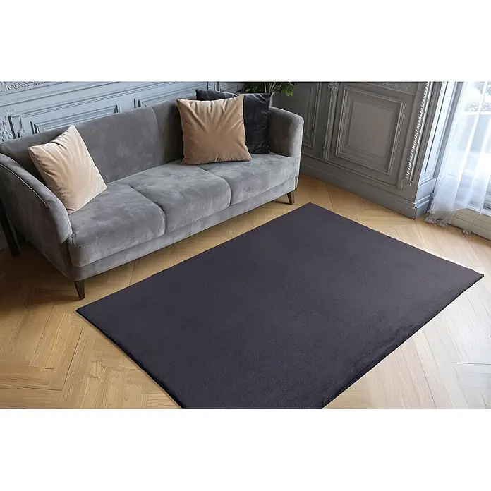 Kayoom Fellteppich Boundless 225 - Anthrazit, 170 x 120 cm, 90 % Polyester, 10 % Baumwolle - Dunkelgrauer Plüschteppich auf hellem Holzboden, vor einem grauen Sofa in einem Wohnzimmer.