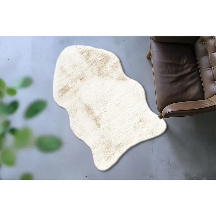 Kayoom Kunstfell Boundless 226 - Creme, 90 x 60 cm, 90 % Polyester, 10 % Baumwolle - Weicher cremefarbener Kunstfellteppich neben einem braunen Ledersessel.