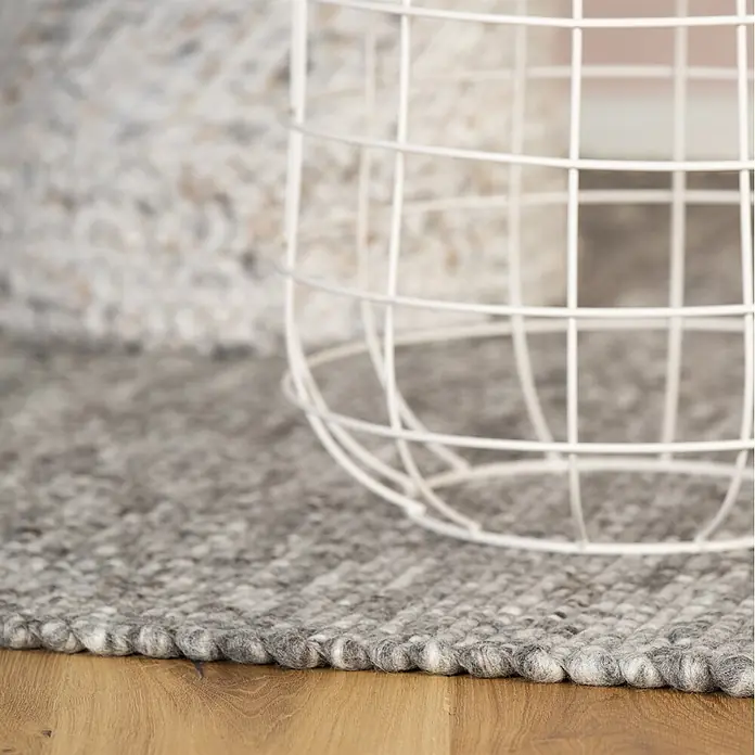 Kayoom Wollteppich Kvell 875 - Silber, 170 x 120 cm, 100 % Wolle - Nahaufnahme eines strukturierten grauen Filz-Wollteppichs auf Holzfußboden, mit teilweise sichtbarem weißen Drahtkorb.