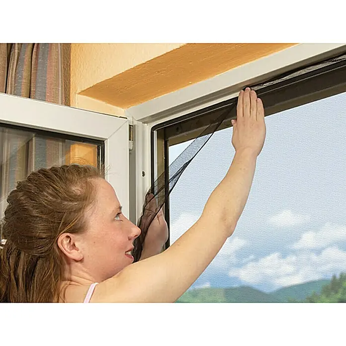 Easy Life Insektenschutzfenster - B x H: 100 x 100 cm, Farbe Netz: Anthrazit, Montage Insektengitter: Ankletten, Kürzbar - Eine Frau befestigt ein Fliegengitter an einem Fenster, um Insekten fernzuhalten und frische Luft hereinzulassen.