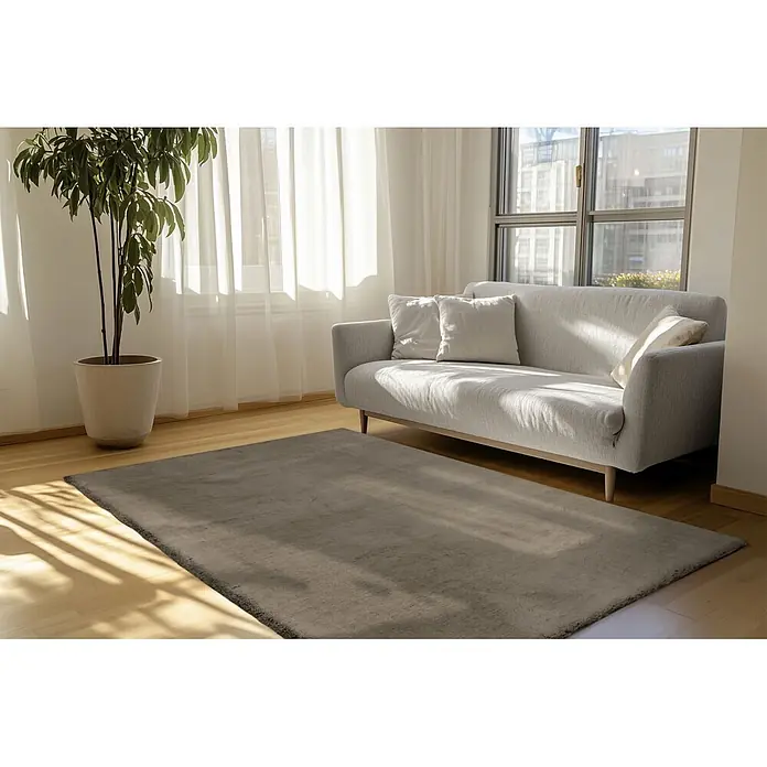 Kayoom Fellteppich Luxe 525 - Wallnuss, 170 x 120 cm, 90 % Polyester, 10 % Wolle - Ein rechteckiger grauer Teppich mit weicher Textur liegt auf einem hellen Holzfußboden in einem Wohnzimmer, teilweise unter einem hellgrauen Sofa.