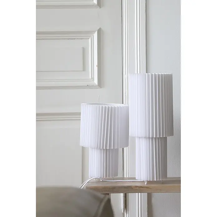 Markslöjd Tischleuchte Catis - L x B x H: 21,5 x 21,5 x 32 cm, E27 - Zwei weiße plissierte Tischlampen auf einem Holzregal in einem hellen Raum.
