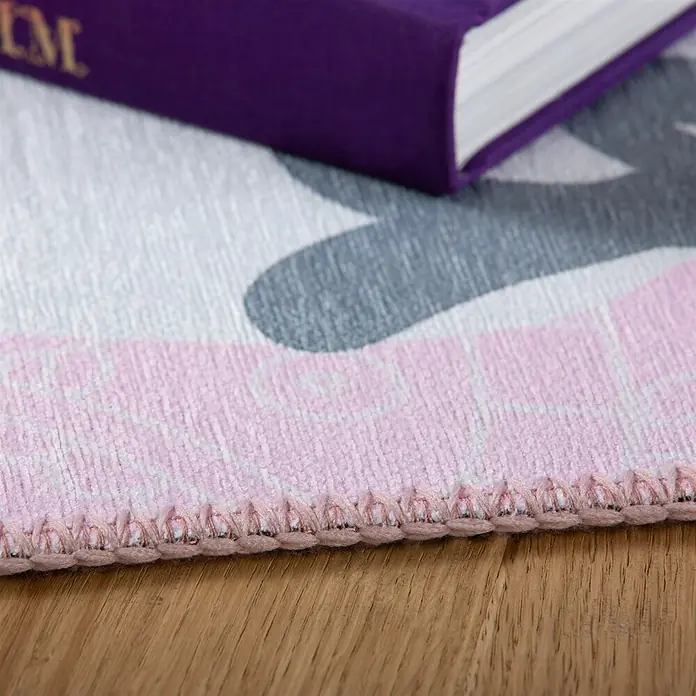 Kayoom Kinderteppich Sympathy Kids 245 White Horse - Weiß, 170 x 120 cm, 100 % Polyester - Nahaufnahme eines rosa, weiß und grau gemusterten Teppichs mit Zierstichrand auf Holzfußboden, mit teilweise sichtbarem lila Buch.