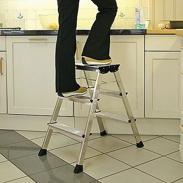 Krause Monto Escalerilla plegable - Número de peldaños: 6 ud., Altura de trabajo: 2,65 m, Carga soportada: 150 kg, Aluminio - Escalera de 3 peldaños de aluminio plateado, en uso en una cocina, sosteniendo a una persona.