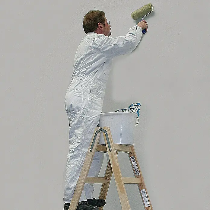 Krause Stabilo Escalera doble - Número de peldaños: 10 ud., Altura de trabajo: 2,75 m, Carga soportada: 150 kg, Madera - Un pintor con mono está de pie en una escalera de madera, utilizando un rodillo de pintura para pintar una pared. Un cubo de pintura en la escalera.