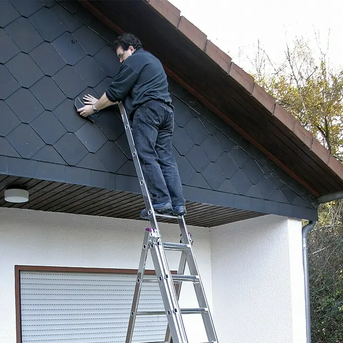 Krause Corda Escalera multiusos - Altura de trabajo: 6,2 m, Número de peldaños: 27 ud., Carga soportada: 150 kg, Aluminio - Hombre en una escalera reparando un techo. La escalera es plateada y está frente a un edificio blanco.