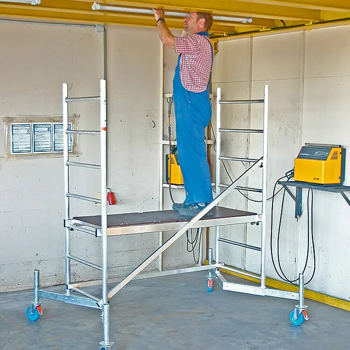Krause ClimTec Andamio móvil - Altura de trabajo: 3 m - Un hombre está de pie sobre un andamio móvil y repara una lámpara de techo. El andamio tiene ruedas y es de aluminio.