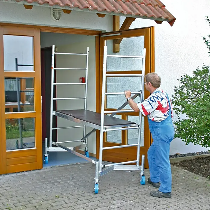 Krause ClimTec Andamio móvil - Altura de trabajo: 3 m - Un hombre empuja un andamio móvil con una plataforma de trabajo frente a una puerta. El andamio tiene ruedas azules y un marco de color plateado.