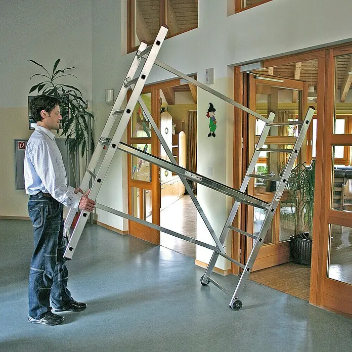 Krause Corda Kleingerüst - Arbeitshöhe: 3 m - Mann transportiert eine vielseitige, zusammenklappbare Aluminium-Mehrzweckleiter mit Rollen in einem Innenraum.