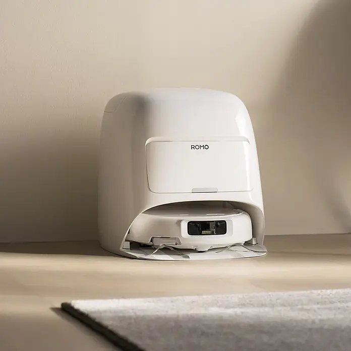 DJI Saugwischroboter S - Saugkraft: 25.000 Pa, Akkulaufzeit: 180 min, Fassungsvermögen Roboter: 260 ml - Weißer ROMO Saugroboter in seiner schlanken Ladestation auf hellem Holzboden.