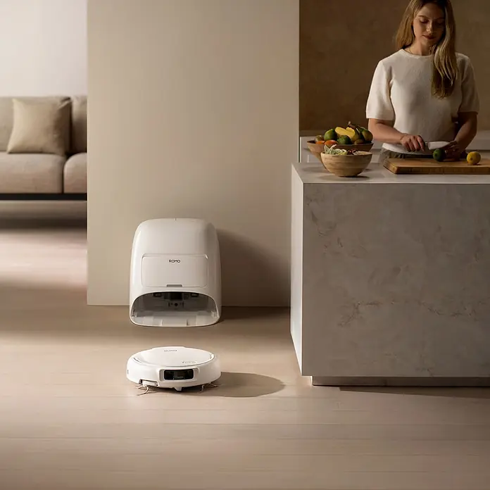 DJI Saugwischroboter S - Saugkraft: 25.000 Pa, Akkulaufzeit: 180 min, Fassungsvermögen Roboter: 260 ml - Weißer Saugroboter und Selbstreinigungsstation auf Holzboden in modernem Zuhause, daneben kochende Frau.
