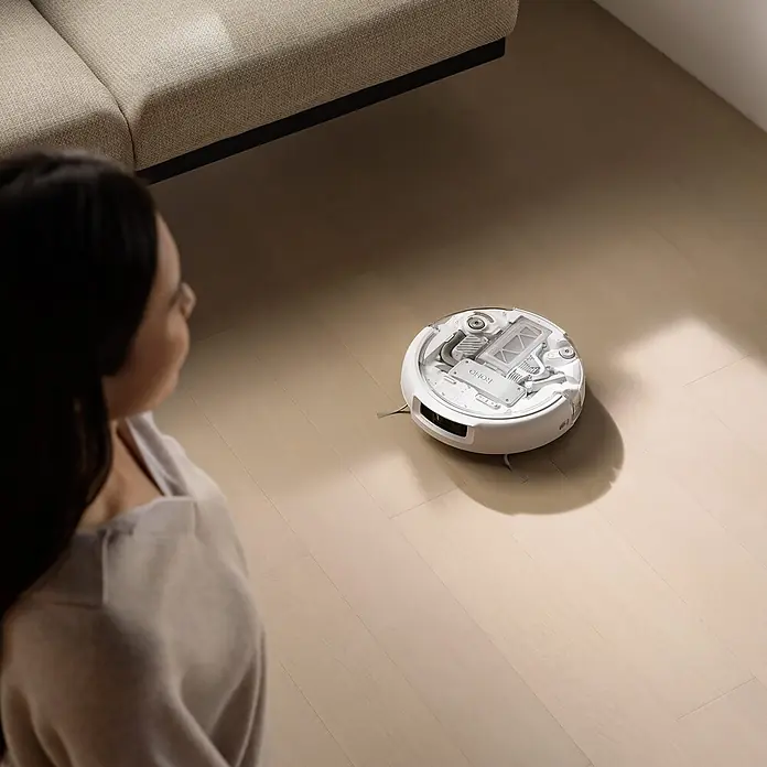 DJI Saugwischroboter A - Saugkraft: 25.000 Pa, Akkulaufzeit: 180 min, Fassungsvermögen Roboter: 260 ml - Weißer Saugroboter mit geöffneter Abdeckung, der interne Komponenten auf einem hellen Holzboden zeigt, von einer Person beobachtet.